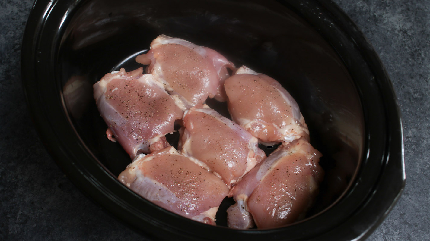 Adding seasoned raw chicken thighs into the bottom of the slow cooker in a single layer.